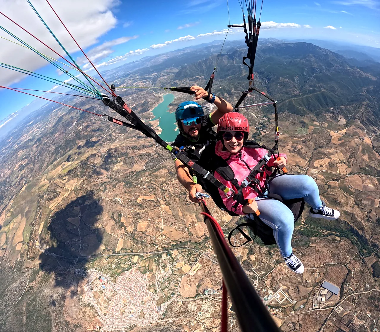 Pareja volando en parapente biplaza sobre un embalse y un pueblo de la sierra